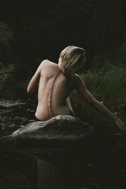 a woman naked in the upper area posing at her back sitting on the stone at the side of a river