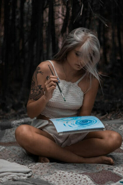 a woman with tattoo on her right arm sitting down on the sand and painting