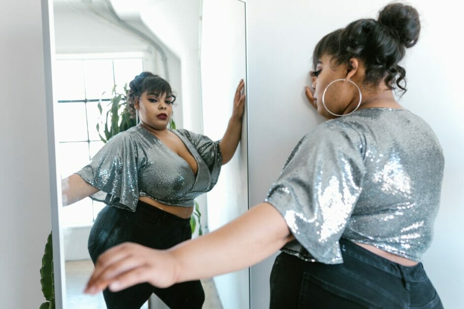 plus-size woman in a silver blouse and black jeans looking at the mirror