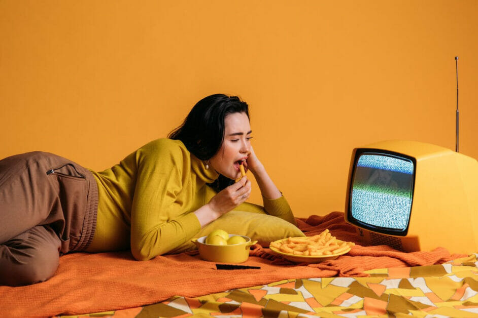woman in a yellow-orange theme photoshoot lying side on the bed with food and tv