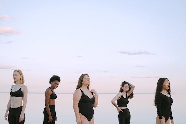 5 women with different body types poses at the beach with their black bikinis/workout dress