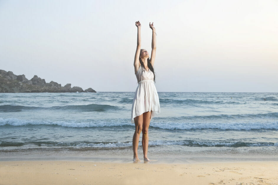 woman in a white dress possess a soft natural kibbe body type lifting her two hands up at the beach